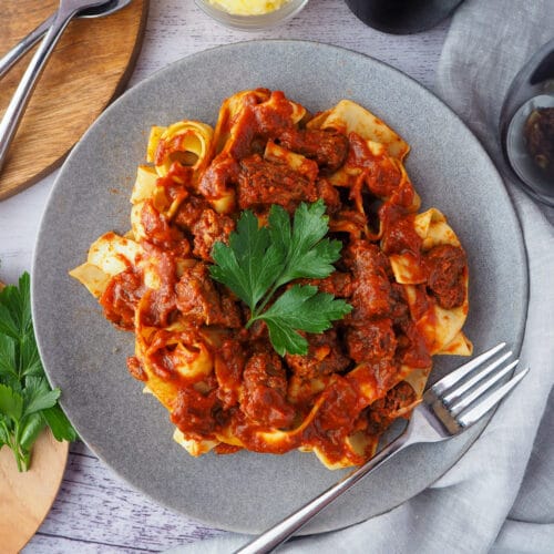 Slow cooker lamb ragu 2 Plate of slow cooker lamb ragu, garnished with parsley, surrounded by serving cutlery, glass of red wine, bowl of parmesan cheese and chopping board with parsley on it.