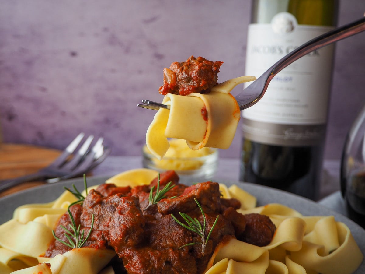 Slow cooker lamb ragu 26 Fork of slow cooker lamb ragu held above a plate of lamb ragu, with a bottle and glass of wine, and bowl of cheese in the background.