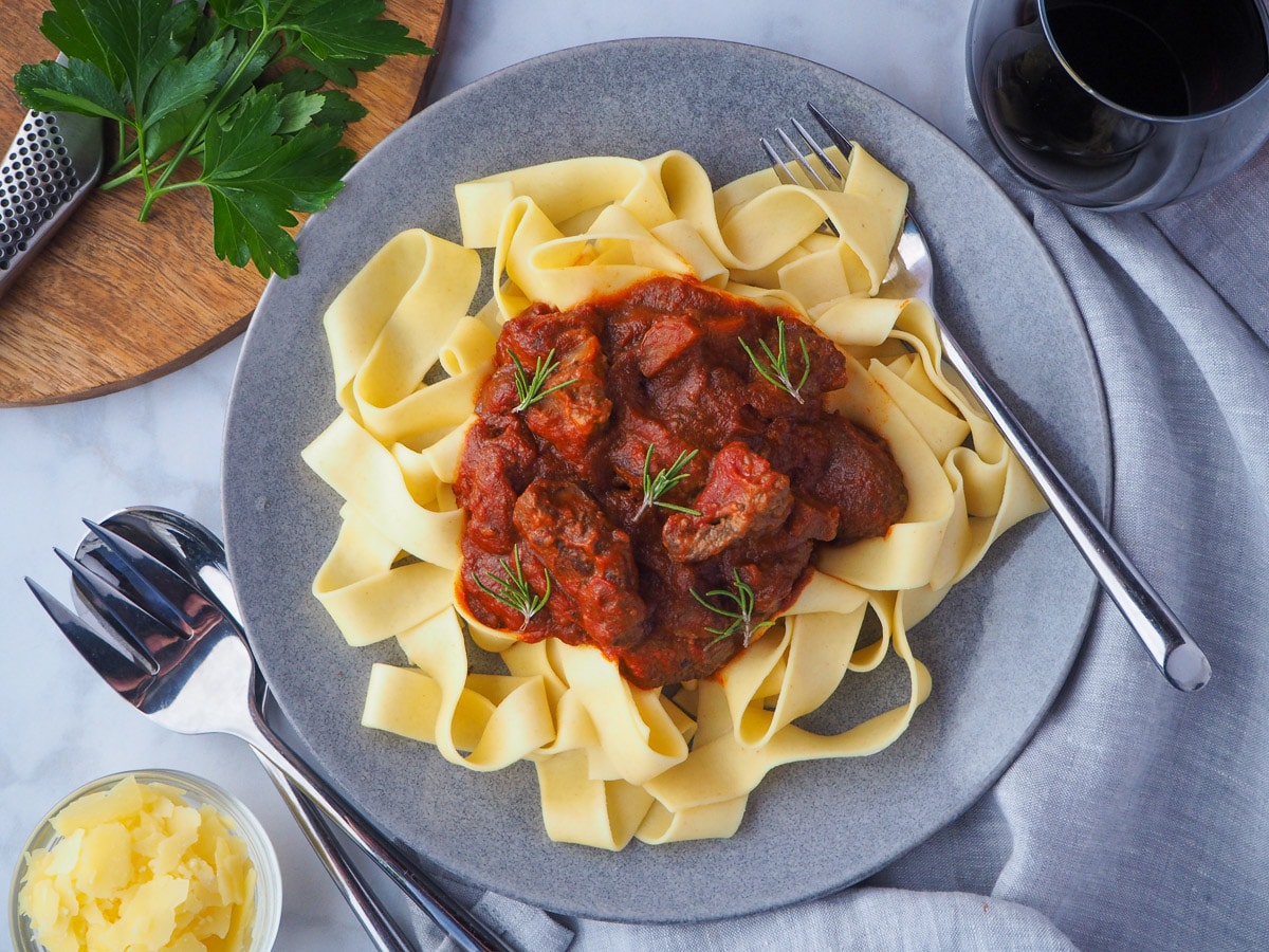 Slow cooker lamb ragu 27 Plate of slow cooker lamb ragu, garnished with rosemary, surrounded by serving cutlery, glass of red wine, bowl of parmesan cheese and chopping board with parsley on it.