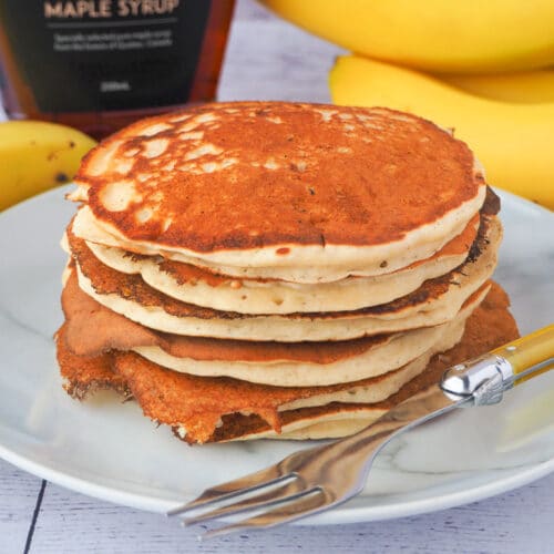 Banana pikelets 1 Close up stack of banana pikelets on a plate, with a vintage spoon on the side, surrounded by fresh whole bananas and bottle of maple syrup in the background.