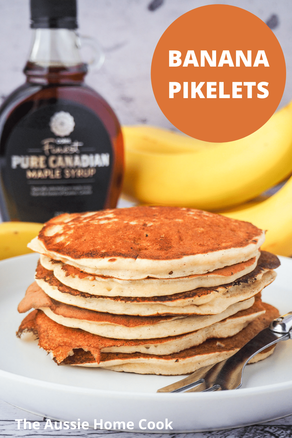 Banana pikelets 17 Stack of banana pikelets on a plate, with a vintage spoon on the side, surrounded by fresh whole bananas and bottle of maple syrup in the background and text overlay, banana pikelets, The Aussie Home Cook.