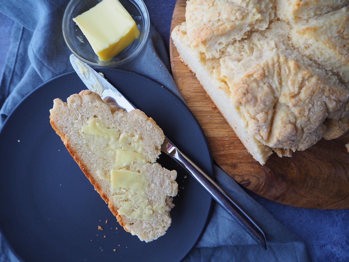 Cut slice of buttered damper on a plate with butter knife, next rest of damper on a chopping board, with butter in dish.