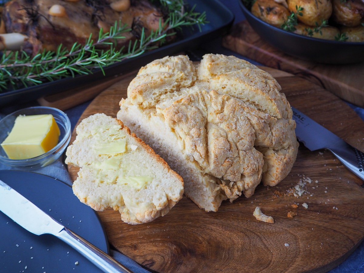 Cut buttered damper on a chopping board, with butter in dish, bread knife and serving dishes with slow cooker lamb shoulder and roasted mini potatoes.