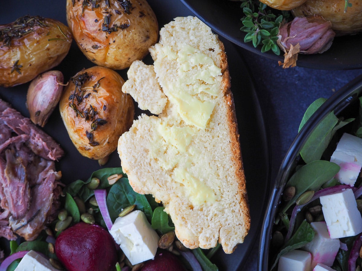 Slice of buttered damper on a plate with beetroot salad, mini roasted potatoes, slow cooker lamb shoulder and larger dishes on the side.