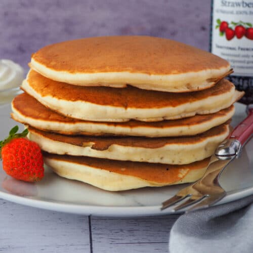 Pikelets 10 Close up stack of pikelets on a plate, with a fresh strawberry and fork, and whipped cream and jar of strawberry jam in the background,