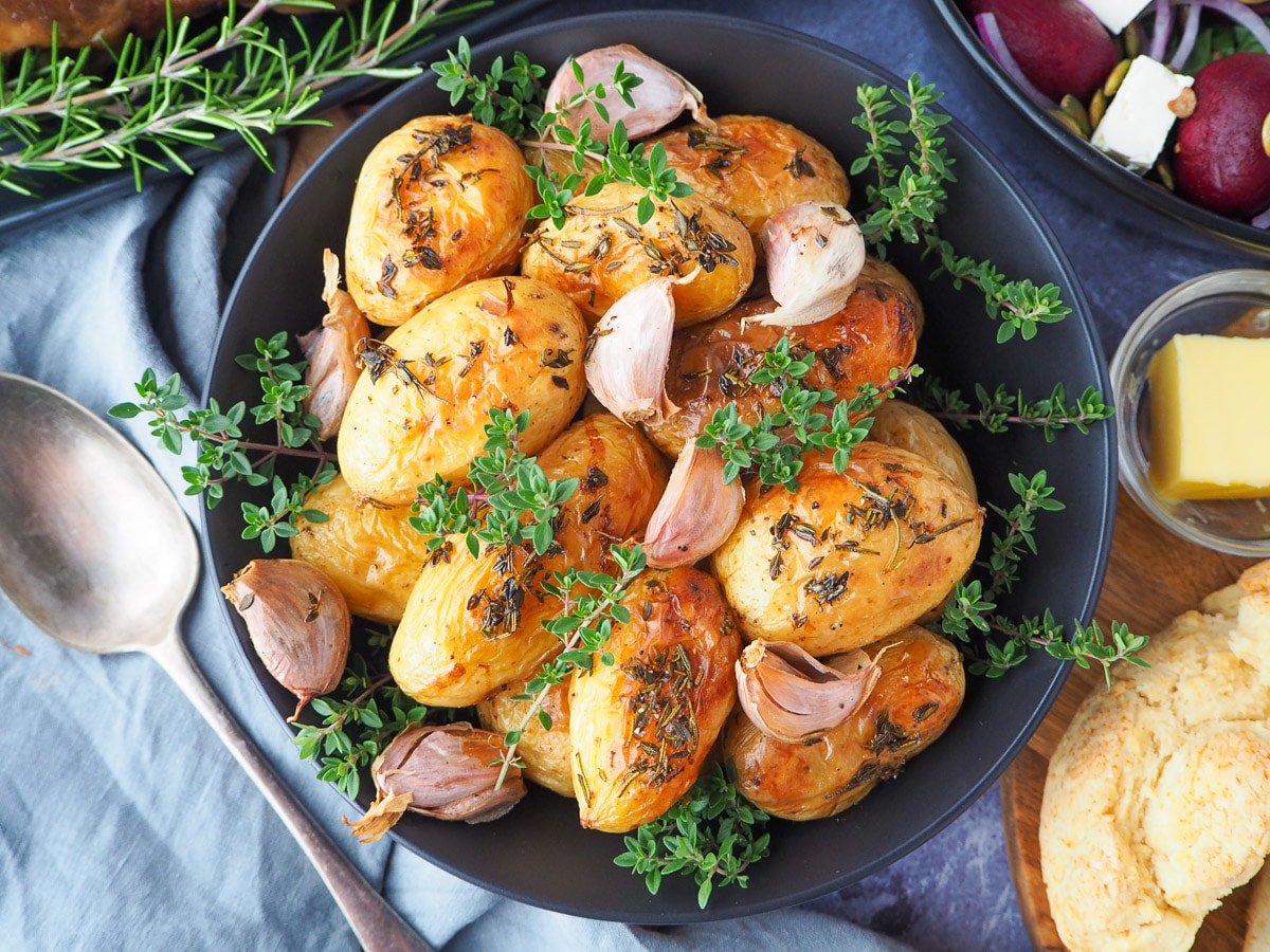 Roasted mini potatoes 1 Roasted mini potatoes in a serving dish garnished with fresh herbs, silver serving spoon to the side, fresh damper, pat of butter, beetroot salad and slow cooker roast lamb on the side.