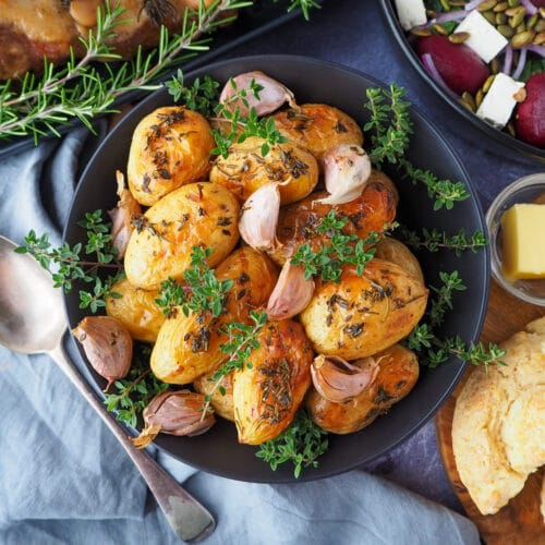 Roasted mini potatoes in a serving dish garnished with fresh herbs, silver serving spoon to the side, fresh damper, pat of butter, beetroot salad and slow cooker roast lamb on the side.