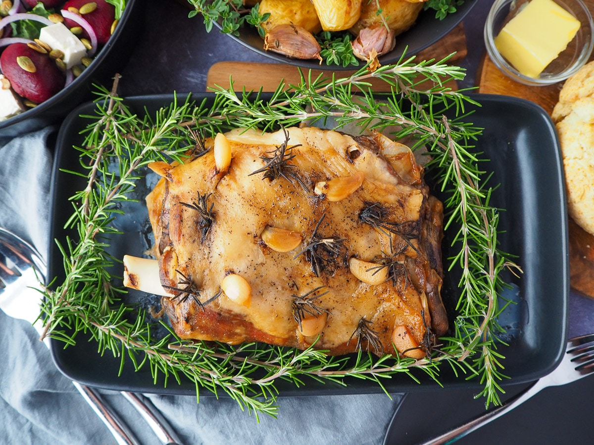 Slow cooker lamb roast on a serving plate, garnished with fresh rosemary, with damper, beetroot salad and roasted mini potatoes on the side.