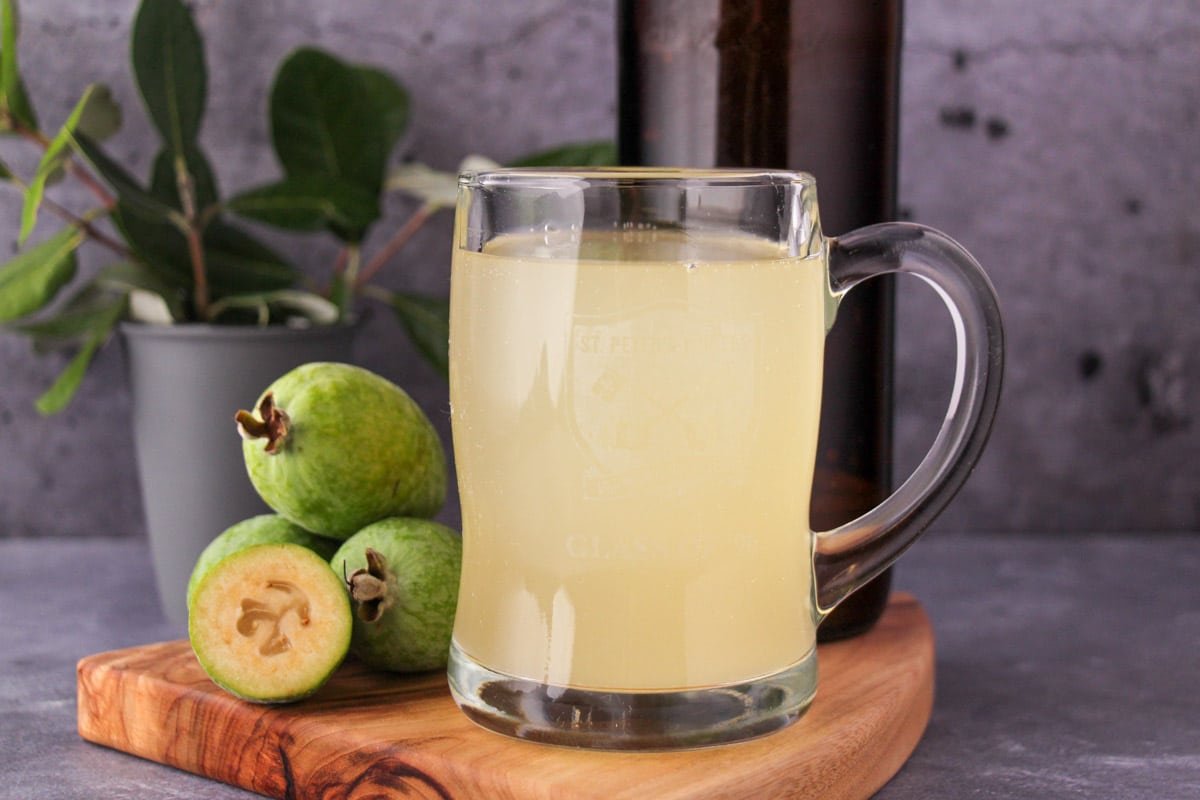 Tankard glass of feijoa fizz on a board, with bottle of feijoa fizz, fresh feijoas and feijoa leaves.