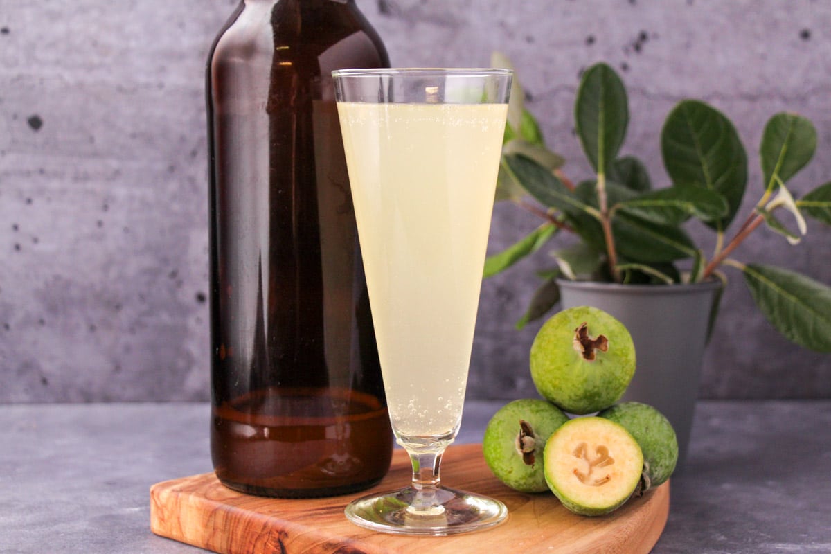 Tall glass of feijoa fizz on a board, with bottle of feijoa fizz, fresh feijoas and feijoa leaves.