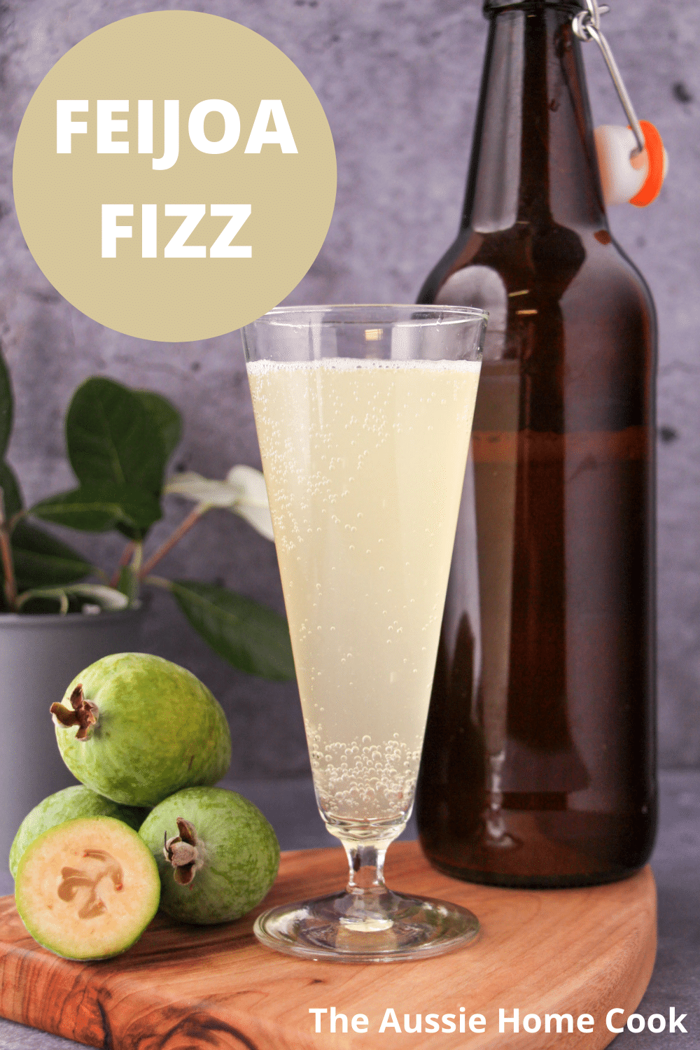 Tall glass of feijoa fizz on a board, with bottle of feijoa fizz, fresh feijoas and feijoa leaves, with text overlay, feijoa fizz and The Aussie Home Cook.