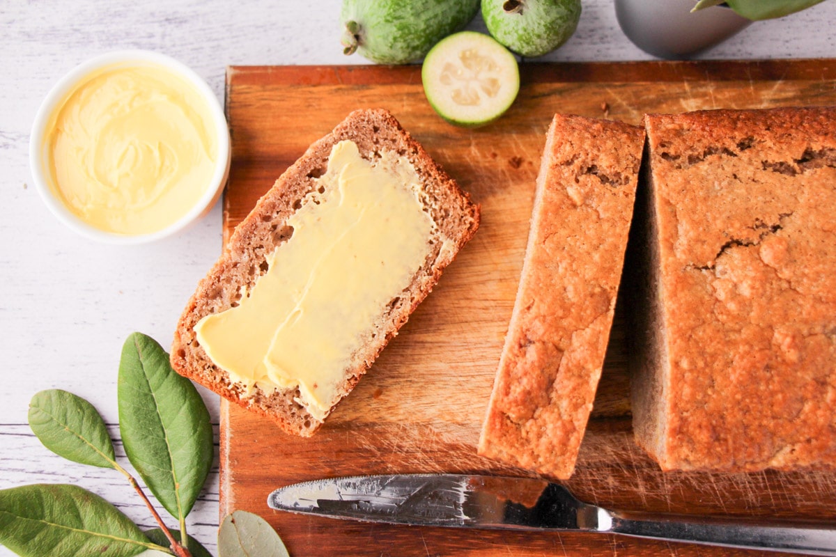 Sliced feijoa loaf on a chopping board, with one buttered slice, fresh feijoas and leaves and a butter knife.