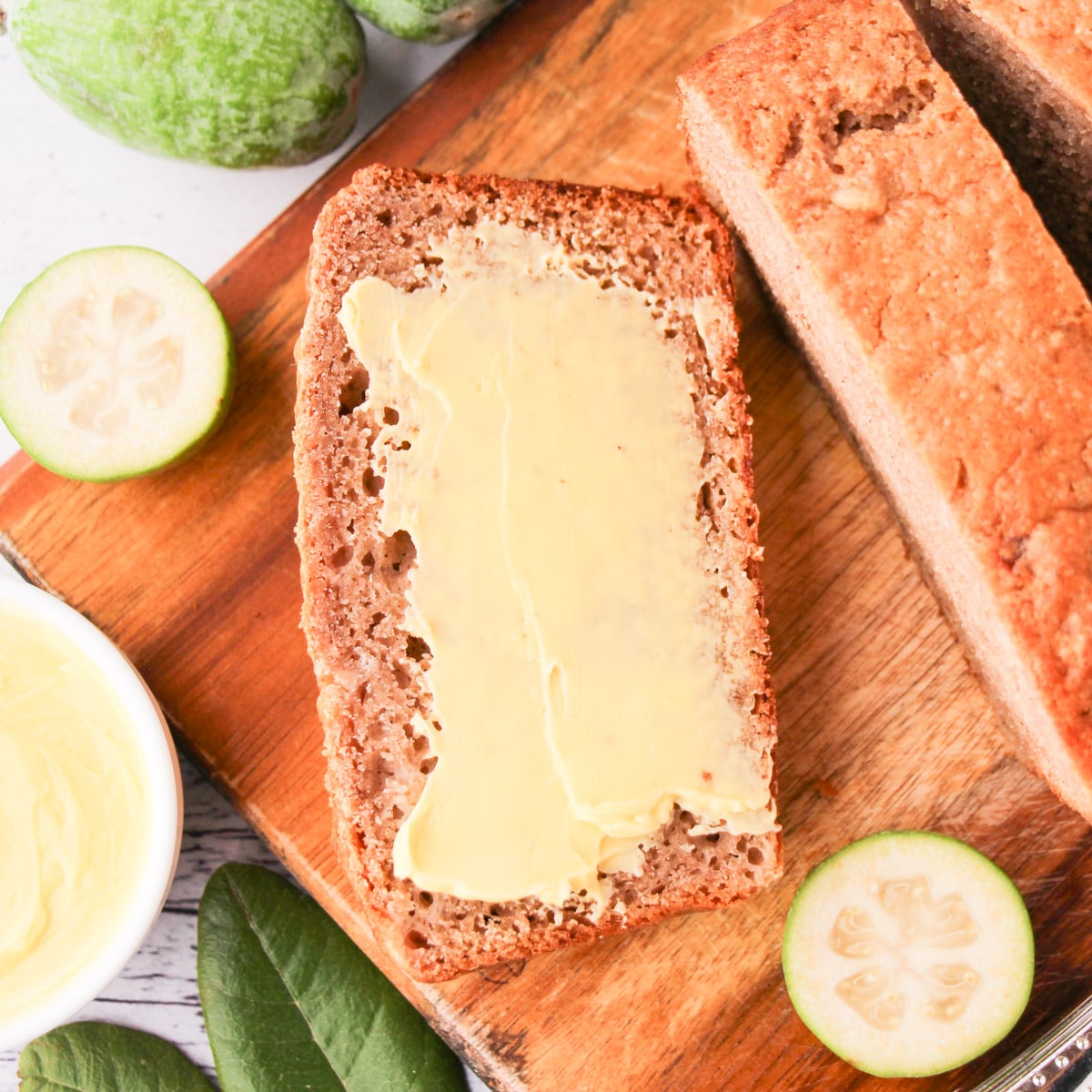 Close up buttered slice of feijoa loaf on a chopping board, with fresh feijoas and leaves and a butter knife.