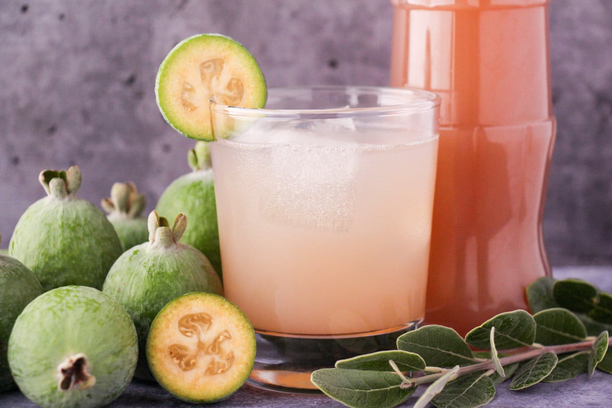 Glass of made up feijoa syrup with ice and fresh feijoa garnish, with bottle of feijoa syrup and fresh feijoas and leaves.
