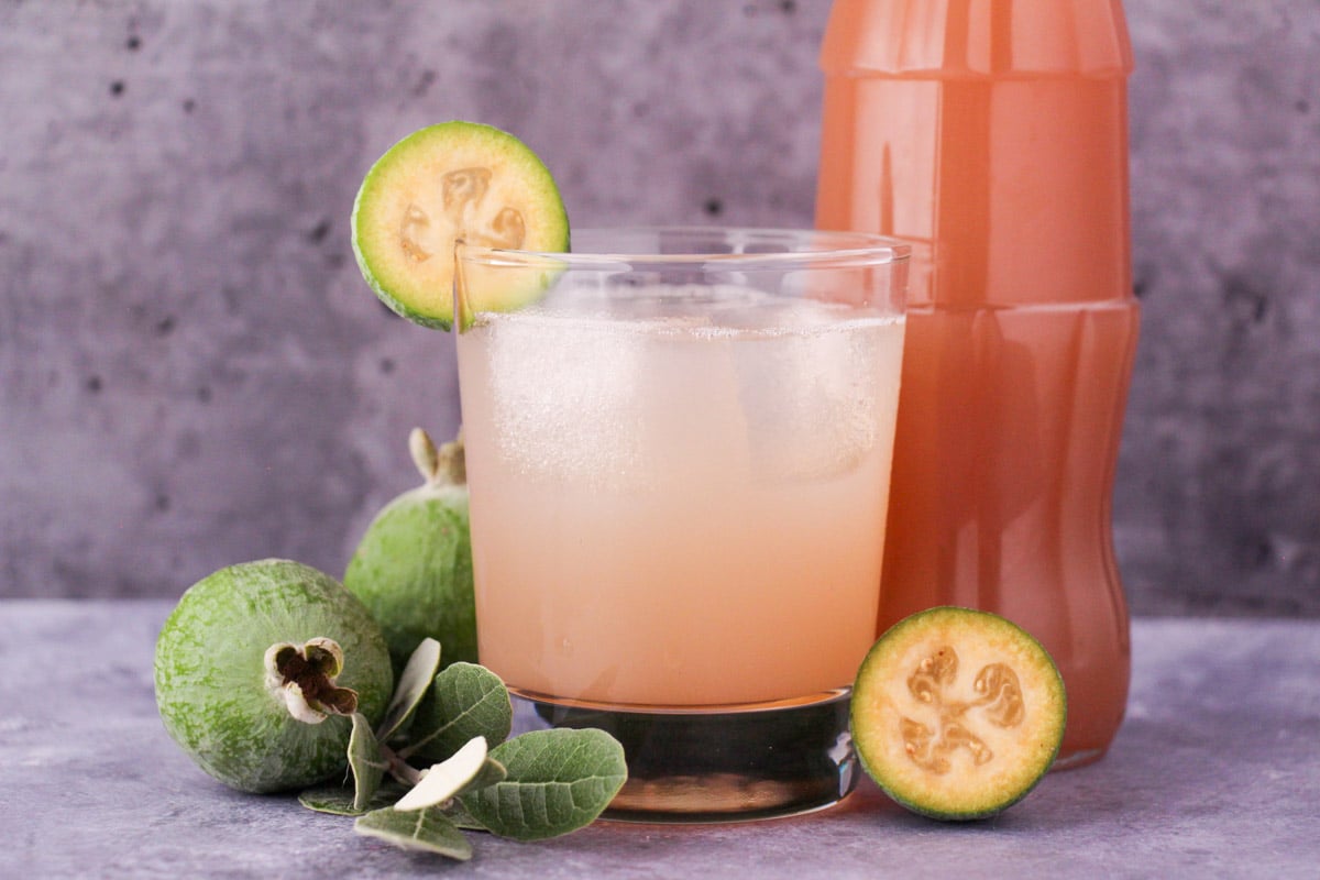 Glass of made up feijoa syrup with ice and fresh feijoa garnish, with bottle of feijoa syrup and fresh feijoas and leaves.