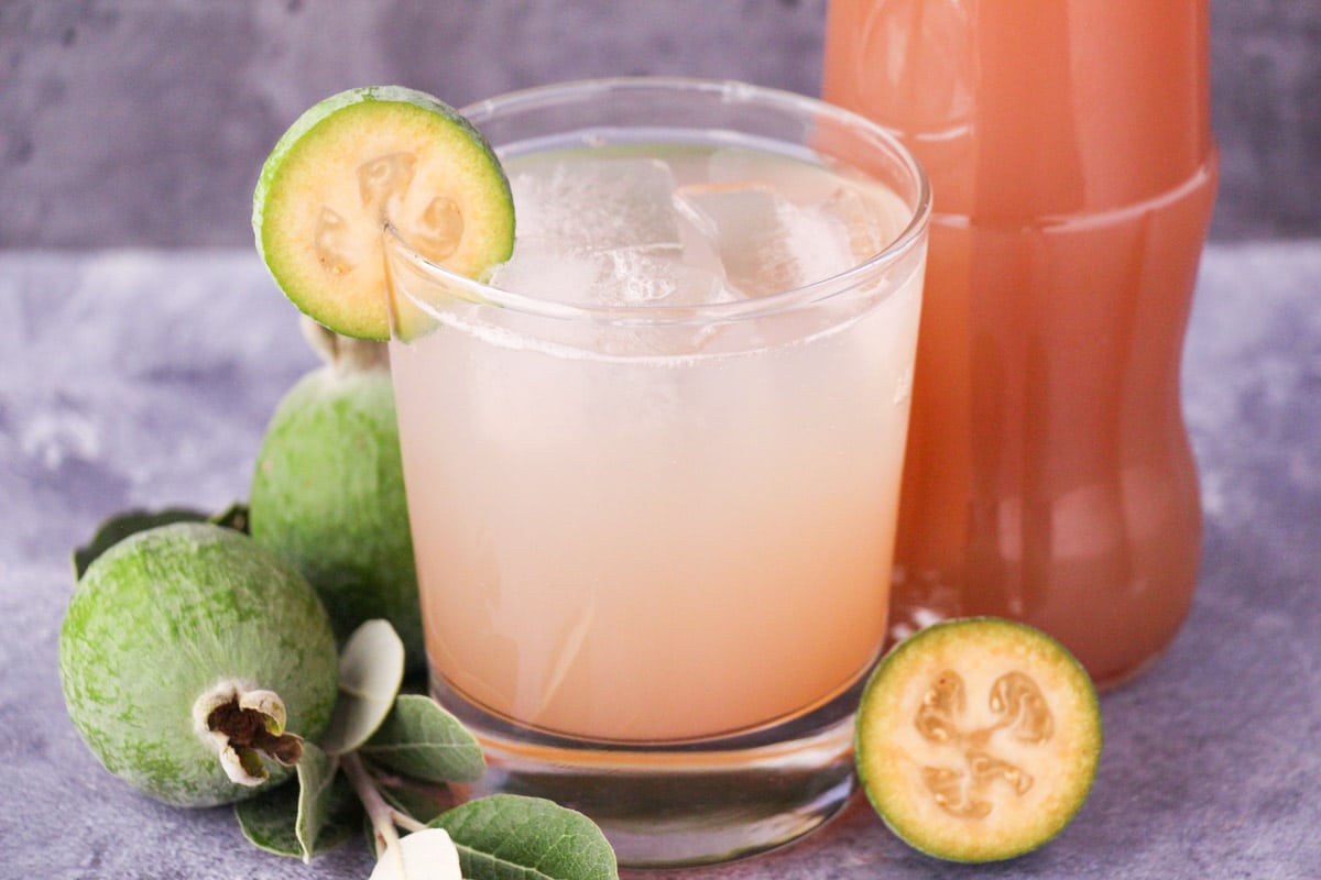 Glass of made up feijoa syrup with ice and fresh feijoa garnish, with bottle of feijoa syrup and fresh feijoas and leaves.