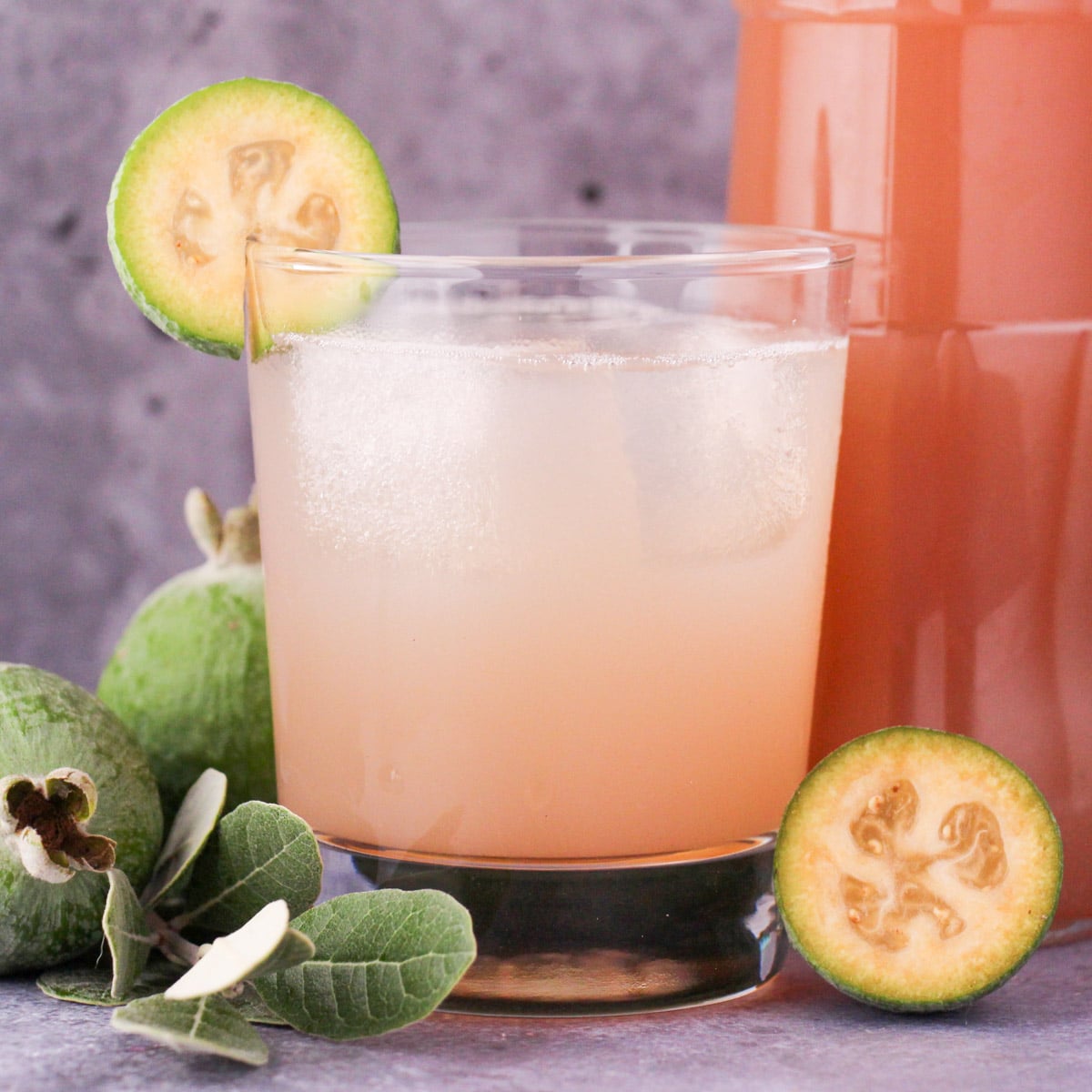 Close up glass of made up feijoa syrup with ice and fresh feijoa garnish, with bottle of feijoa syrup and fresh feijoas and leaves.