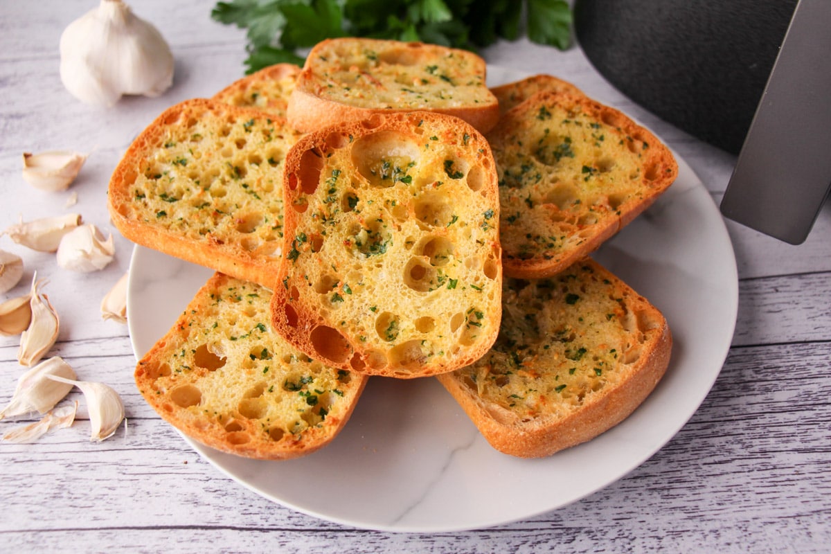 Stack of air fryer garlic bread on a plate, with air fryer basket and fresh garlic cloves to the sides.