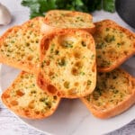Close up of stack of air fryer garlic bread on a plate, with air fryer basket and fresh garlic cloves to the sides.