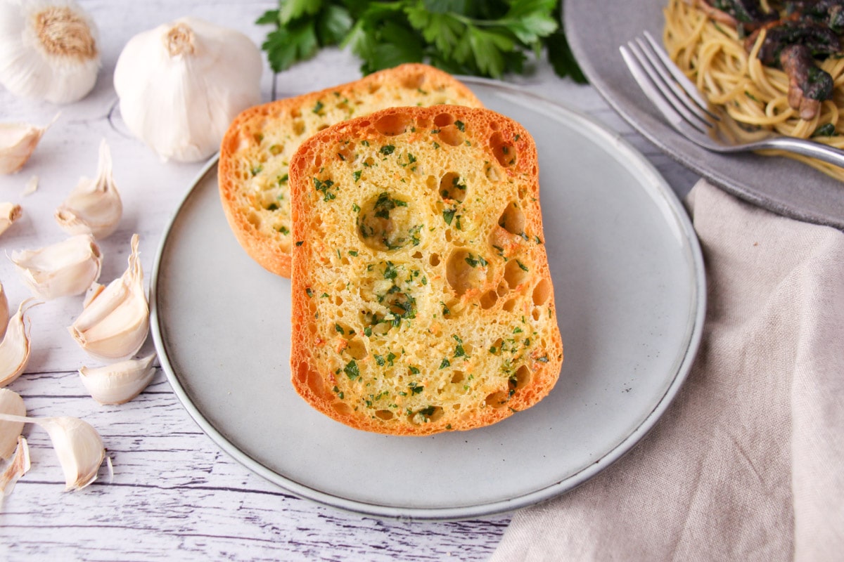 Air fryer garlic bread on a plate, with fresh garlic cloves, fresh parsley and a plate of pasta on the sides.
