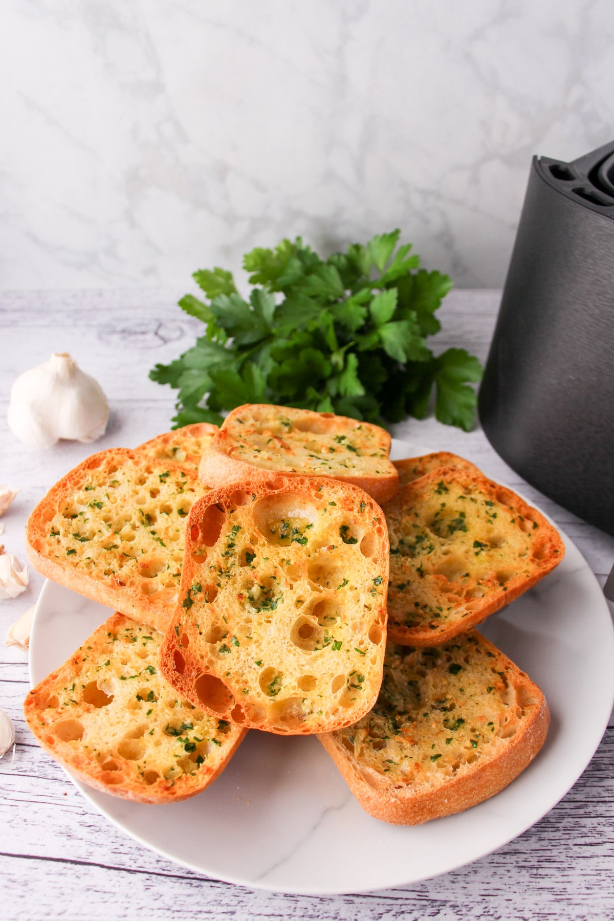 Stack of air fryer garlic bread on a plate, with air fryer basket and fresh garlic cloves to the sides.