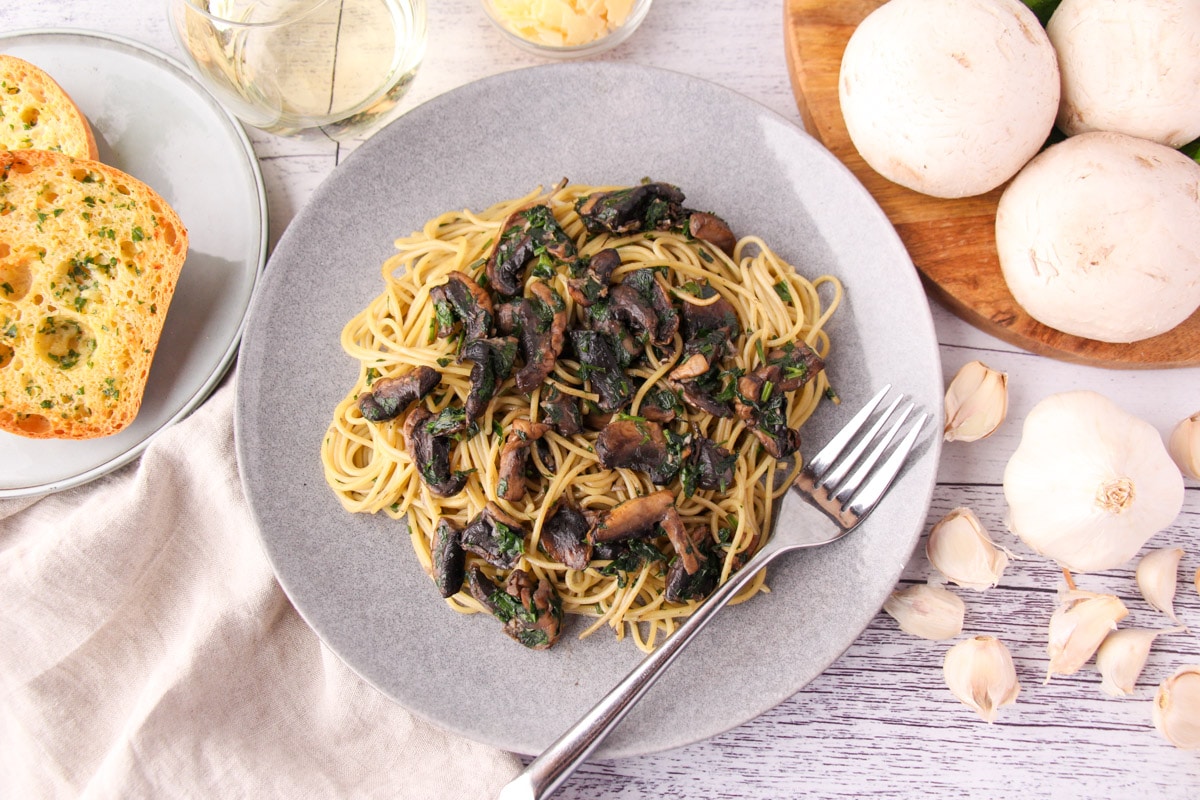 Mushroom aglio olio on a plate with a fork, with garlic bread, fresh garlic, mushrooms and parsley, a glass of white wine and shaved parmesan around the edges.