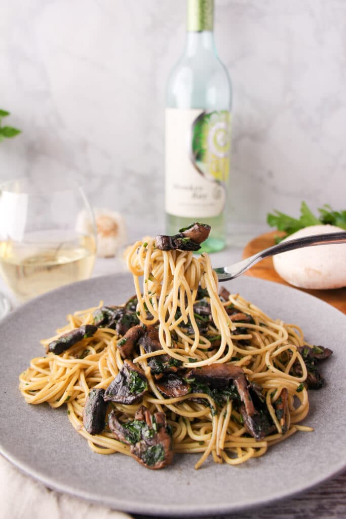90 Dirt cheap meals 22 Fork of mushroom aglio olio held over a plate of pasta, with fresh mushrooms, parsley, garlic and a glass and bottle of white wine the background.