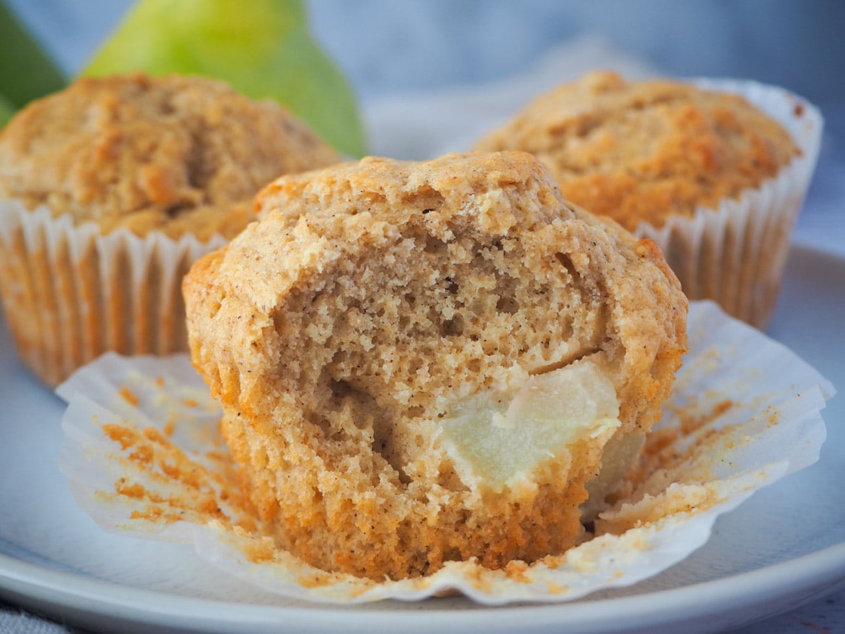 Pear muffins 13 Pear muffin with a piece taken out, with more muffins and fresh pears in the background.