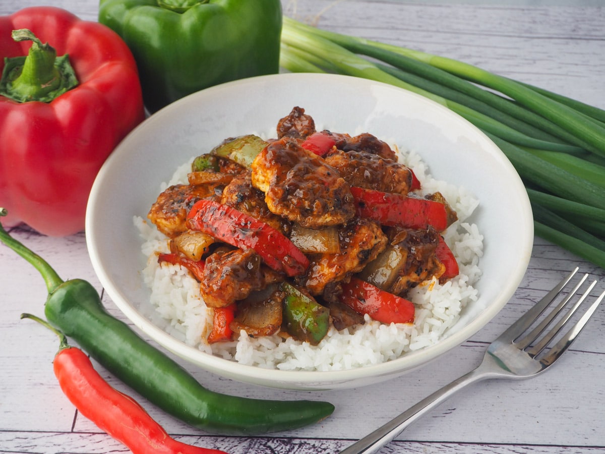 Bowl of chicken on rice with fork and capsicums, spring onions and chilli on the side.