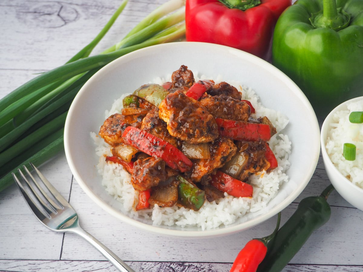 Bowl of chicken on rice with fork, bowl of rice, capsicums, spring onions and chilli on the side.