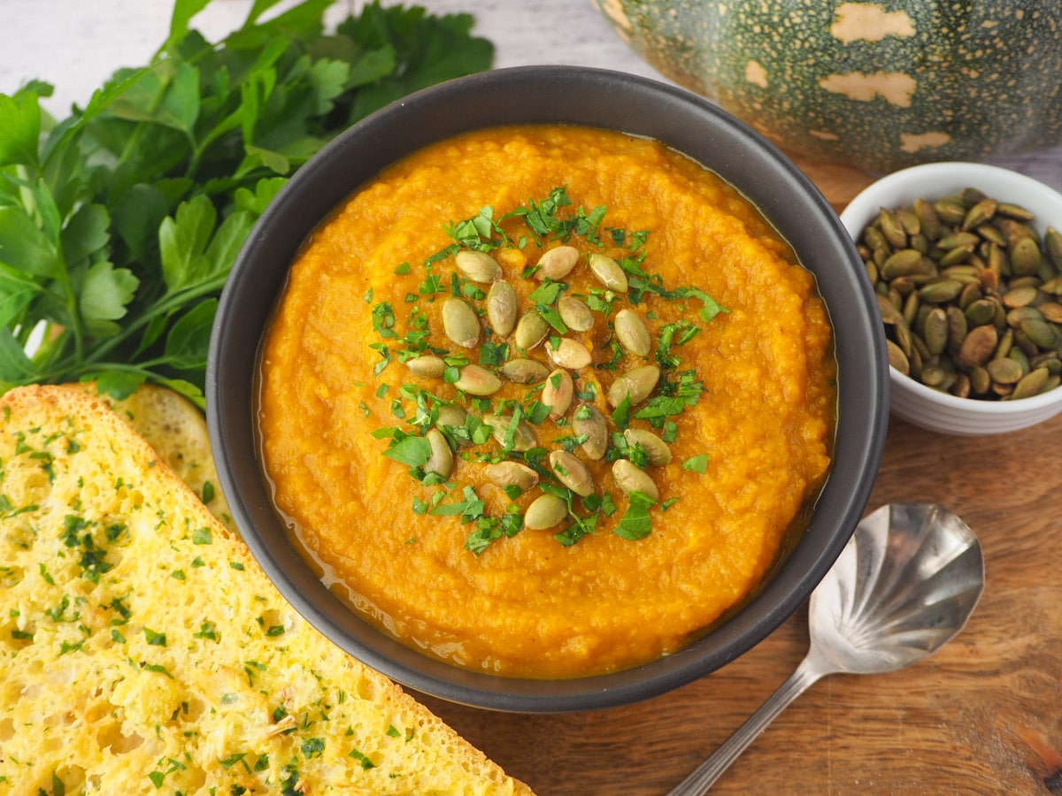 Pumpkin and Sweet Potato Soup 1 Soup with pumpkin seed and parsley garnish, with garlic bread, spoon and toasted pumpkin seeds on the side.