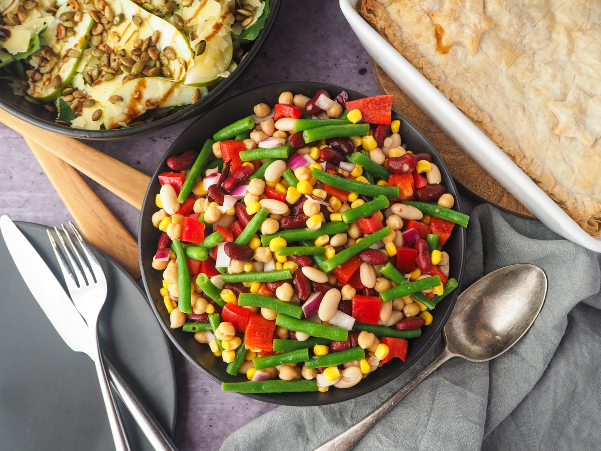 Four Bean Salad 1 Serving bowl of salad with serving spoon, knife, fork and plate on the and pie and rocket and pear salad in the background.