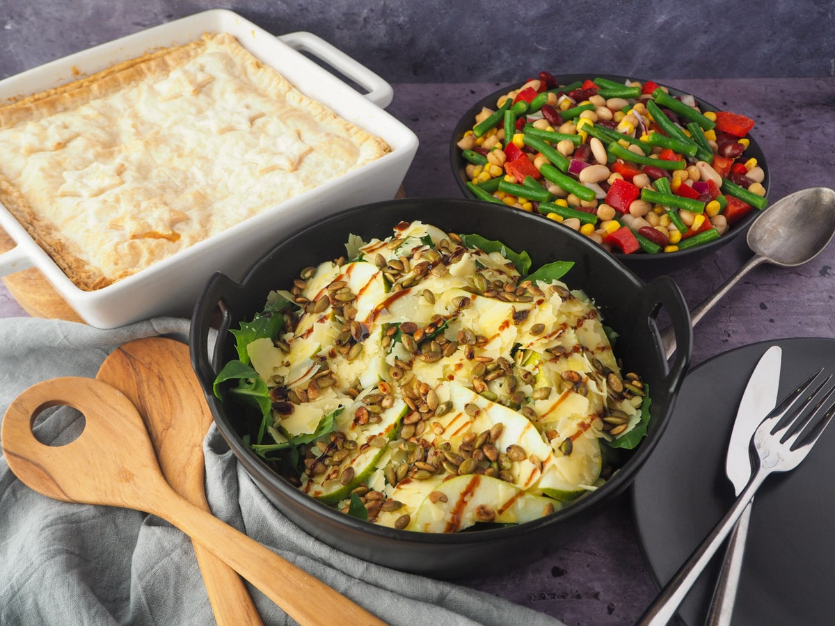 Salad with serving spoons, plate, knife and fork, pie and four bean salad in the background.