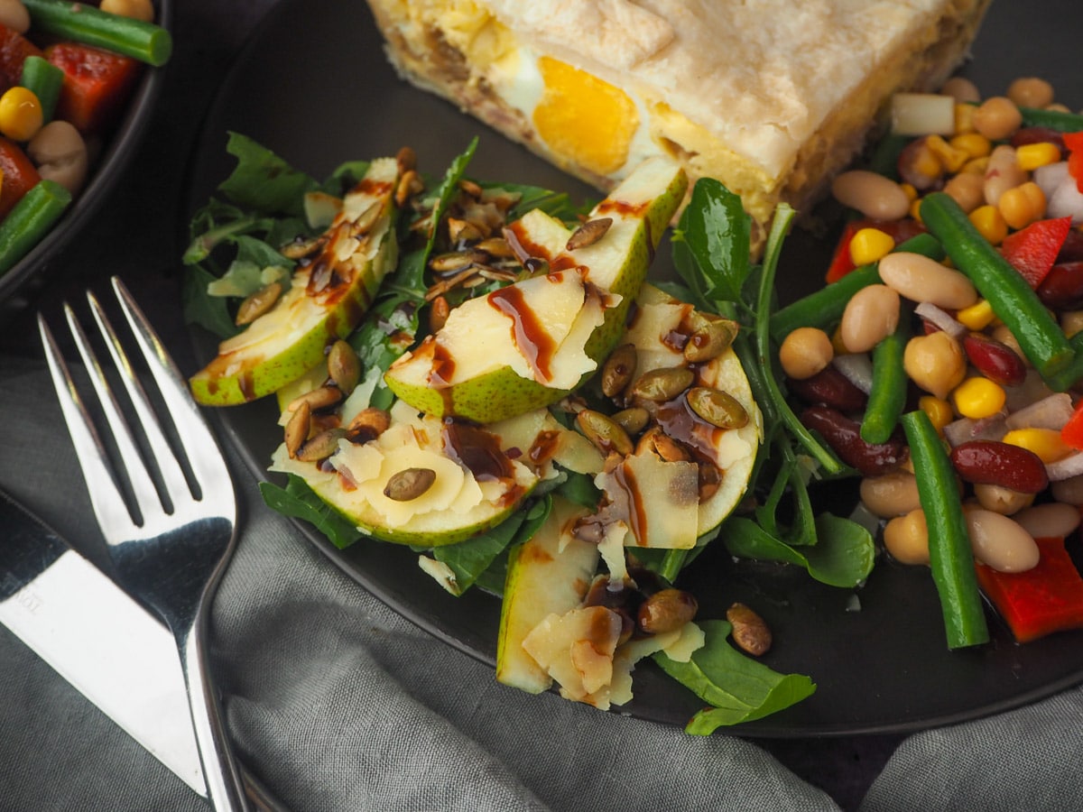 Serving of salad on a plate with bacon and egg pie and four bean salad, knife and fork to the side and four bean salad in the background.