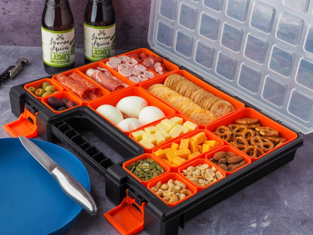 Picnic Charcuterie Board 10 Multi compartment container filled with various charcuterie food, with picnic plate and drinks in the background.