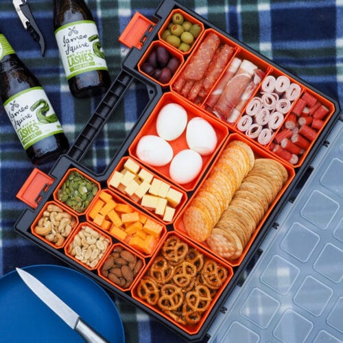 Picnic Charcuterie Board 6 Multi compartment container filled with various charcuterie food, on a picnic blanket with a plate and drinks.