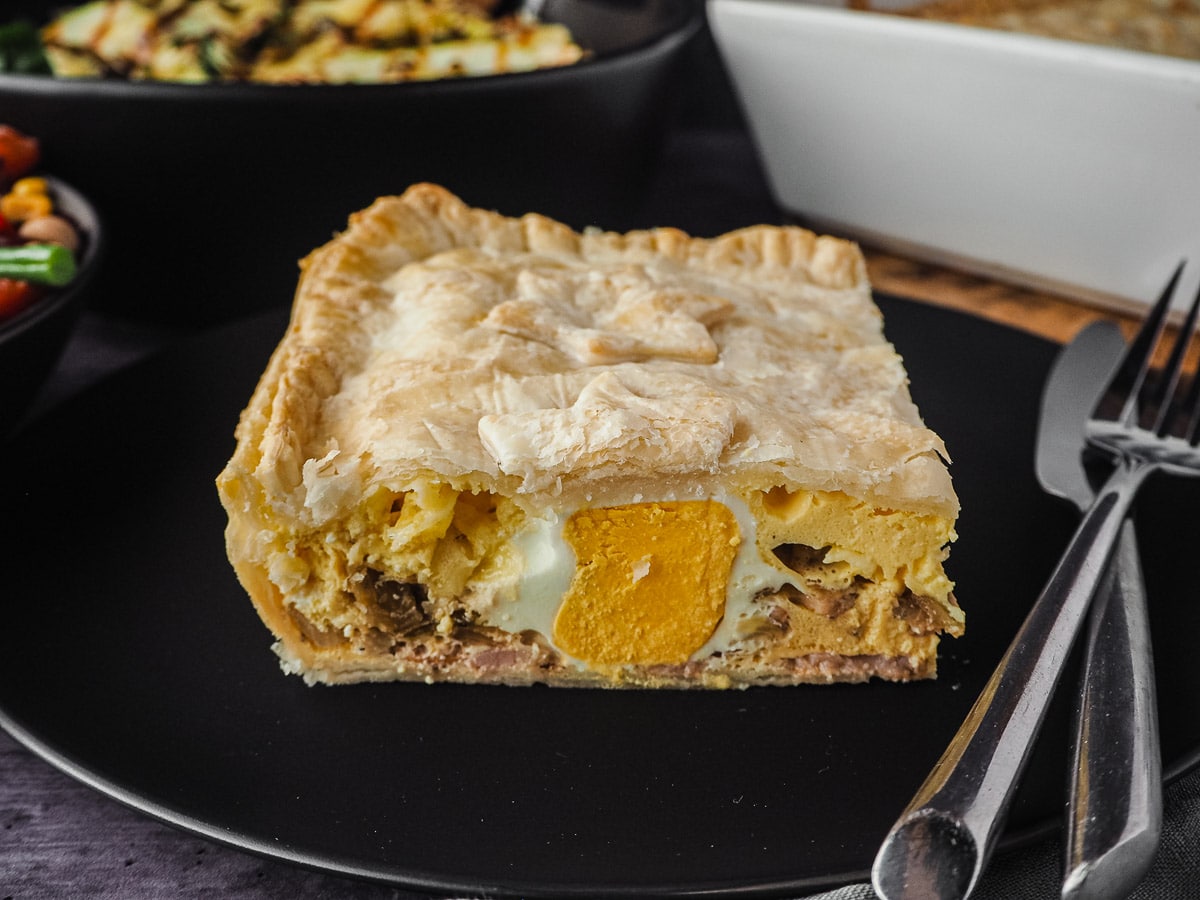 Pie in a plate with cutlery, with pie dish and salads in background.