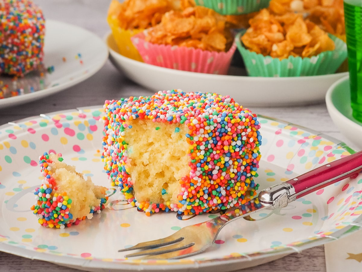 Lamington with one piece out on a plate with a fork and party food in the background.