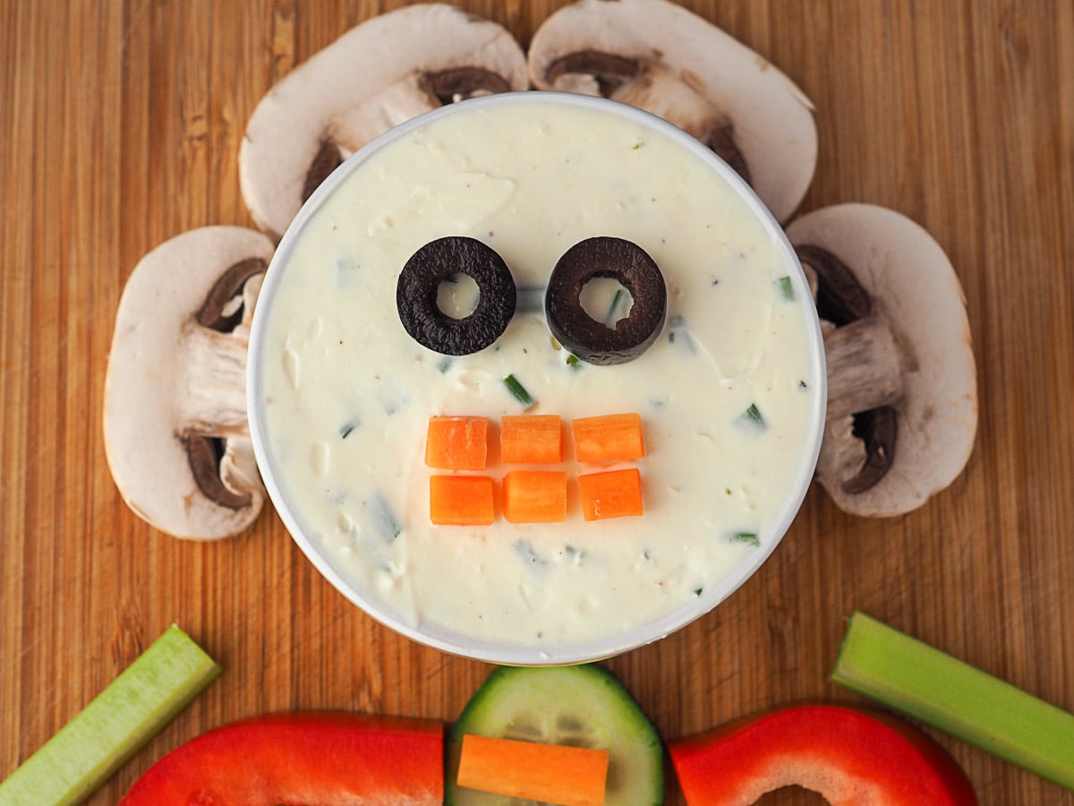 Skeleton Veggie Tray 5 Process shots: dip showing two sliced olive pieces as eyes and carrot cut into teeth shapes. Dip is surrounded by mushroom slices.