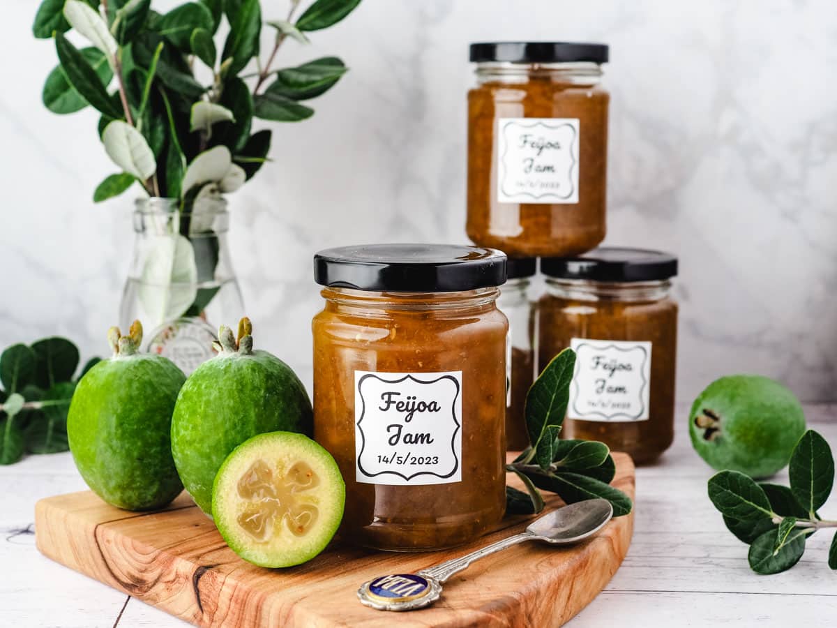 Feijoa Jam 1 Jar of feijoa jam on a serving board with fresh feijoas and leaves on the side, with a stack of feijoa jam jars in the background.