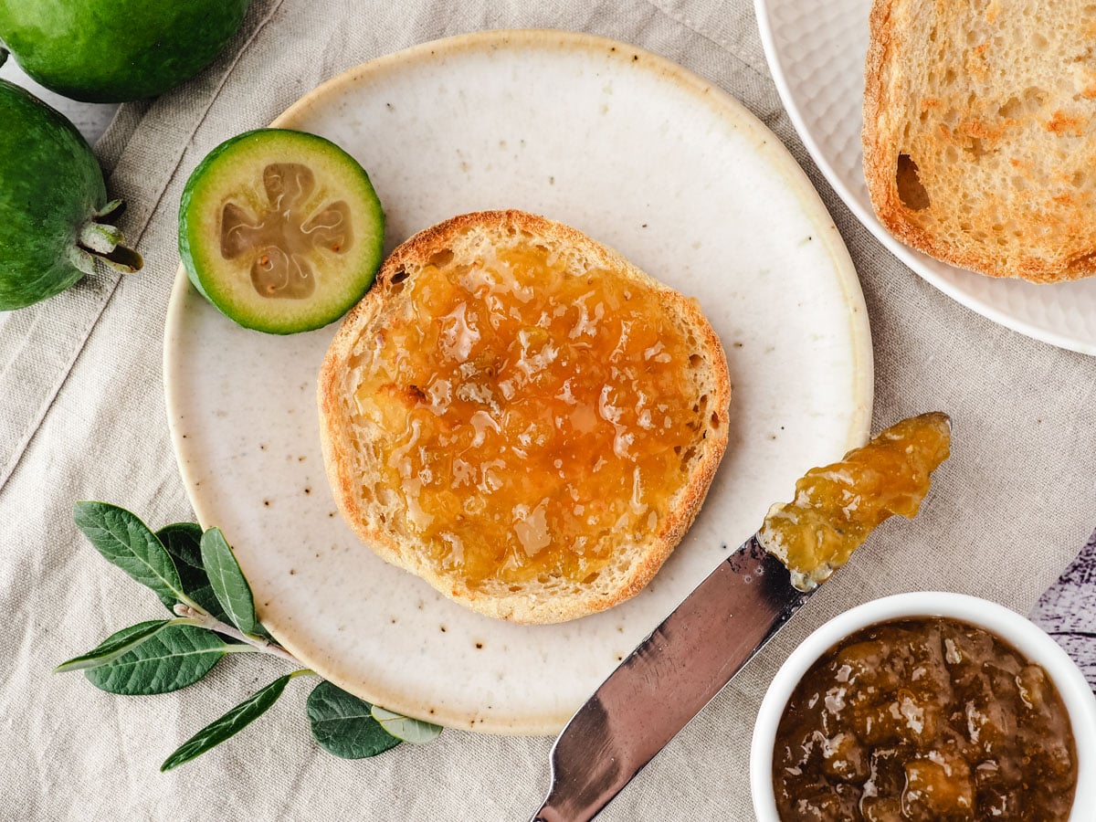 Feijoa Jam 6 Toasted English muffin spread with feijoa jam, on a plate with a knife and fresh feijoas and pot of jam on the side.