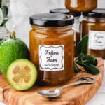 Feijoa Jam 8 Jar of feijoa jam on a serving board with fresh feijoas and leaves on the side, more jars and a plate of toasted muffins in the background.