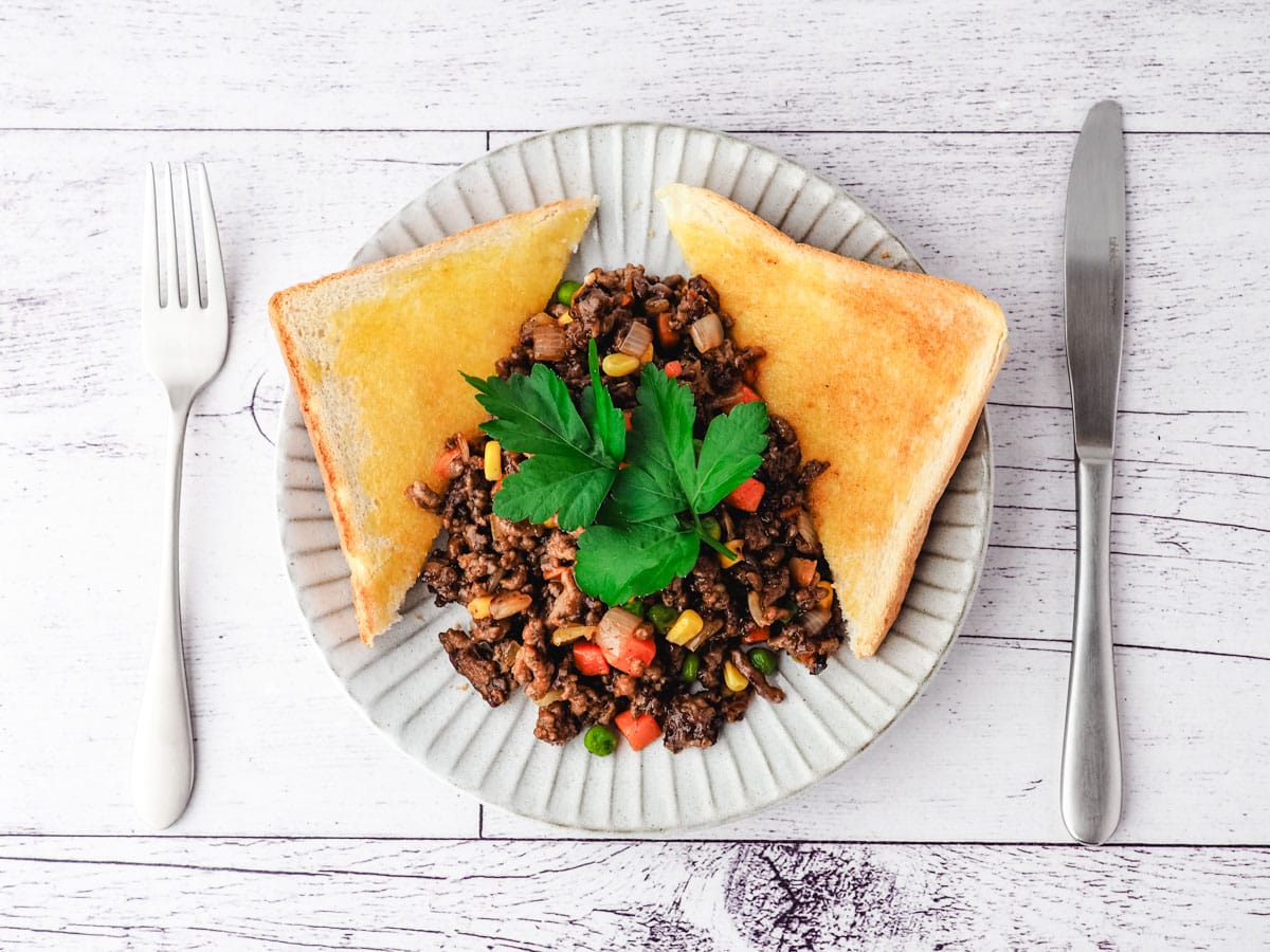 Savory mince on a plate with buttered toast on the side, garnished with flat parsley, with a knife and fork on the side.
