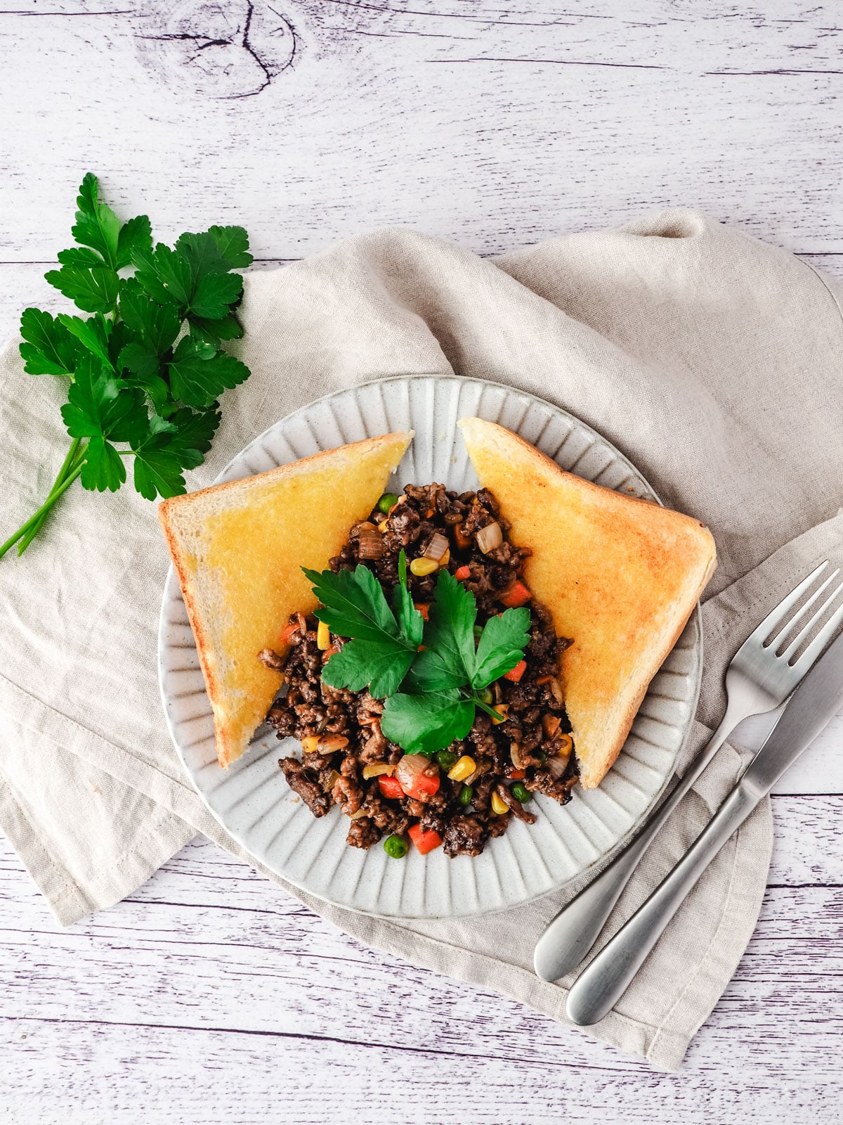 Savory mince on a plate with buttered toast on the side, garnished with flat parsley, with a knife and fork on the side.