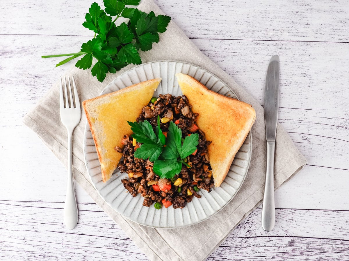 Savory mince on a plate with buttered toast on the side, garnished with flat parsley, with a knife and fork on the side.