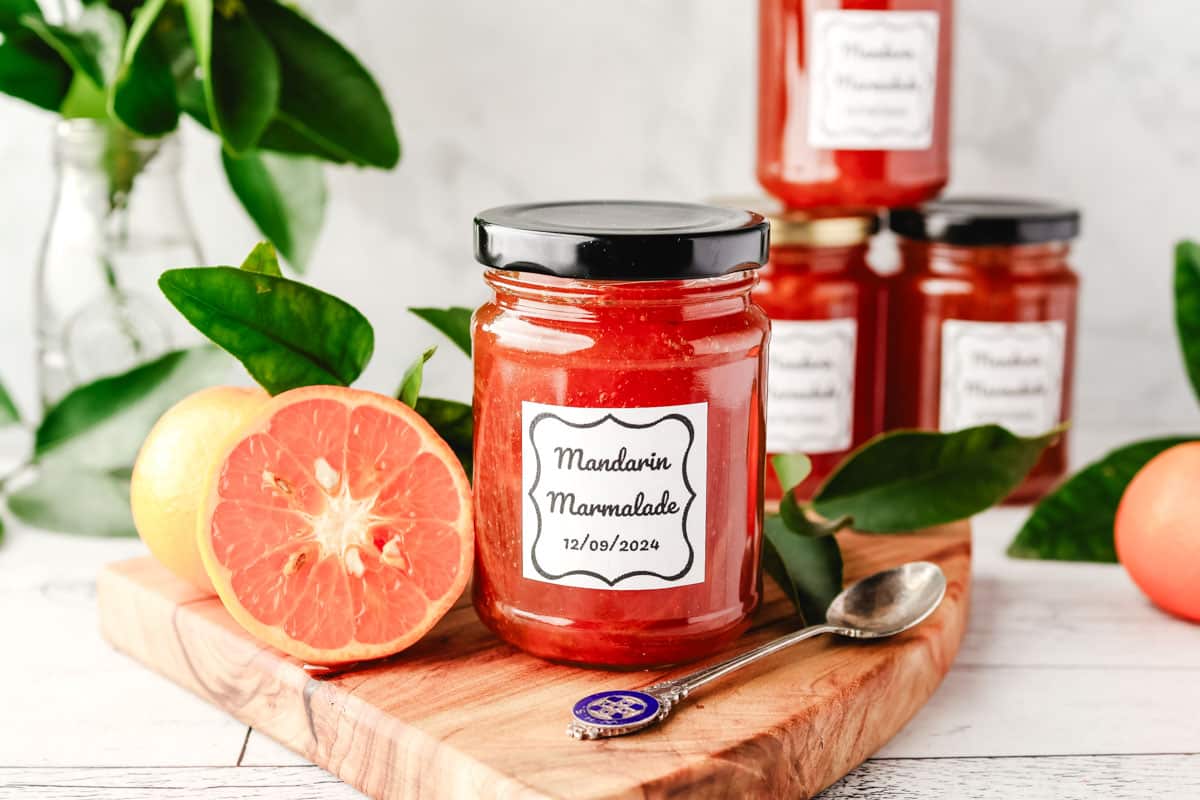 Mandarin Marmalade 1 Jar of mandarin marmalade on a serving board with a spoon, with freshly sliced open mandarin and leaves on the side.