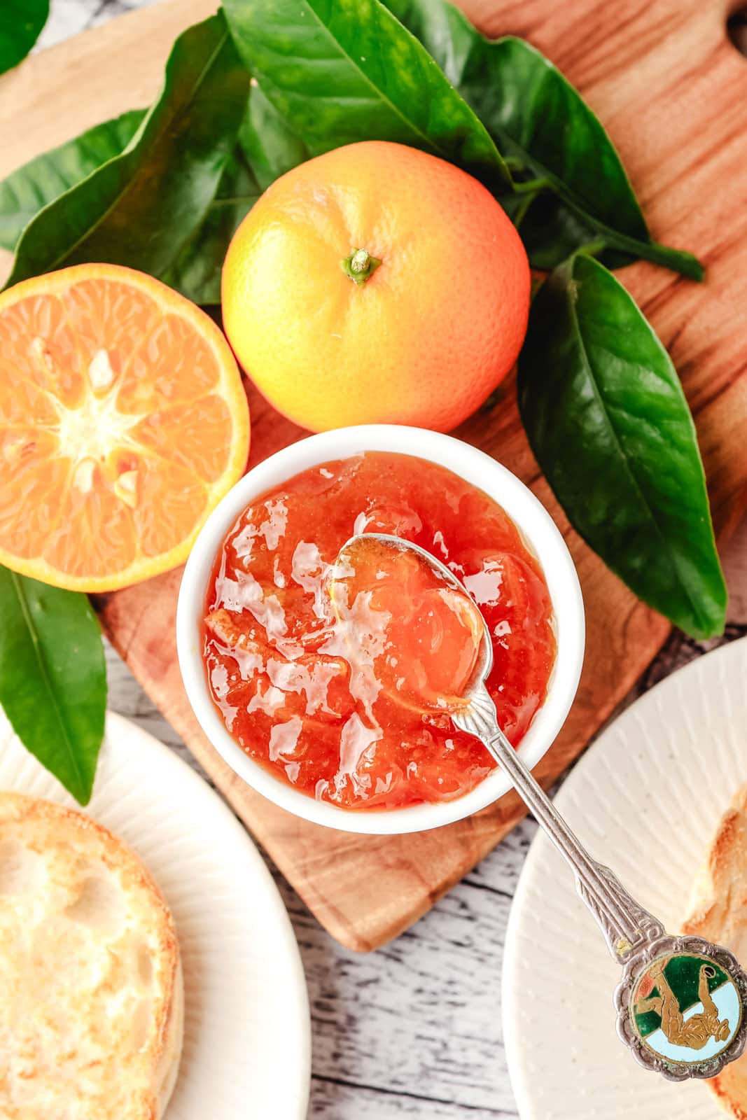 Mandarin Marmalade 8 Top down view of mandarin marmalade in a bowl with a spoon, with toasted English muffins and fresh mandarins on the side.