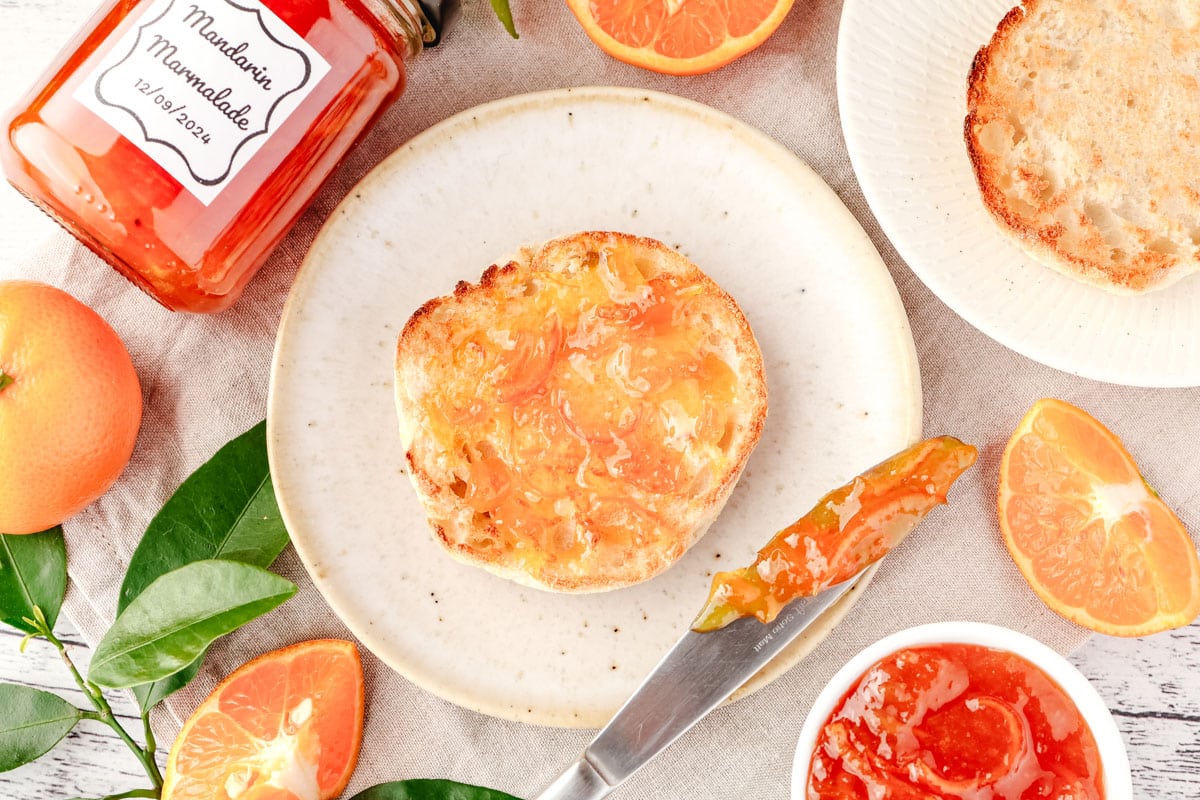 Mandarin Marmalade 5 Top down view of mandarin marmalade on a toasted English muffin, on a plate with serving knife and jar of marmalade on the side.